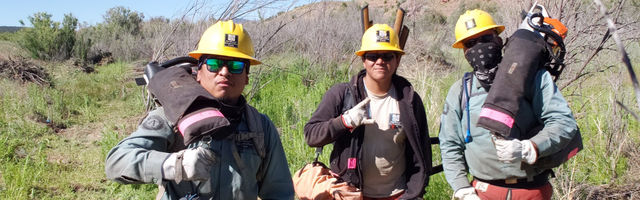 3 members with their gear with grass and mountains in the background